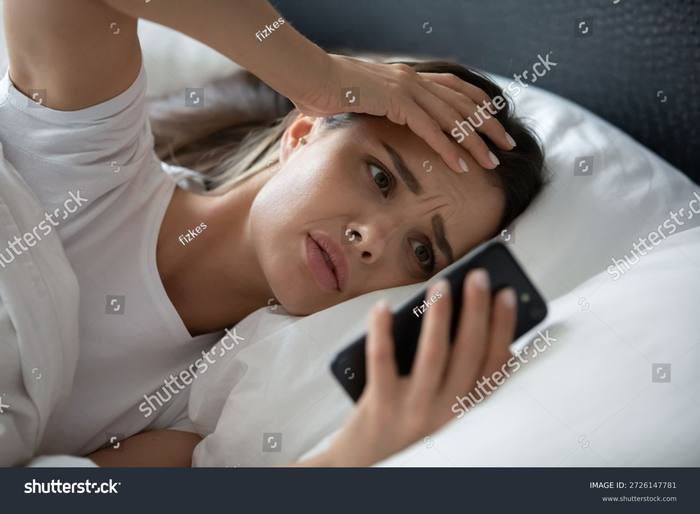 A woman lying awake at night, hand at her forehead, phone in her hand