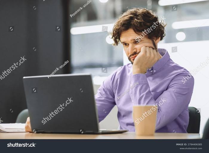 A man at a laptop in an office, chin resting on his hand, looking away from the screen