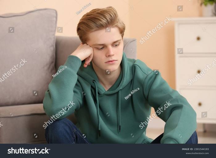 A teenage boy sitting on the floor against a couch, head propped on his hand